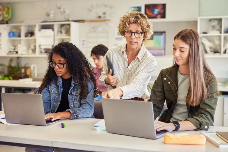 Teacher helps two students as they work on laptops at a classroom desk, guiding their activity.